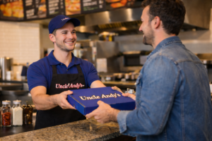 Customer picking up a pizza at Uncle Andy’s Pizza in Farmington MI with employee handing over a blue pizza box at the counter